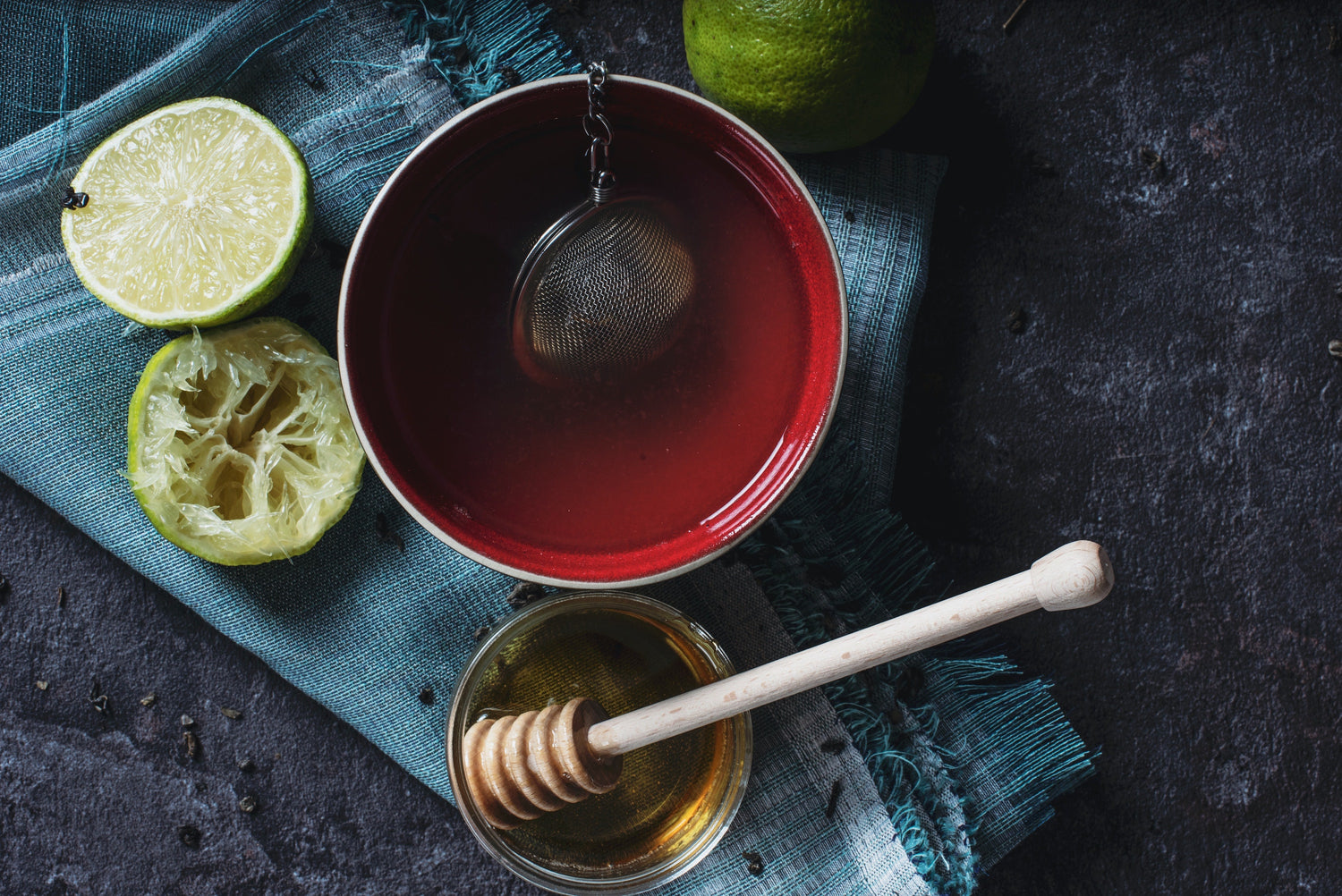 Red bowl of brewed tea with tea strainer, sliced limes, and honey on dark background