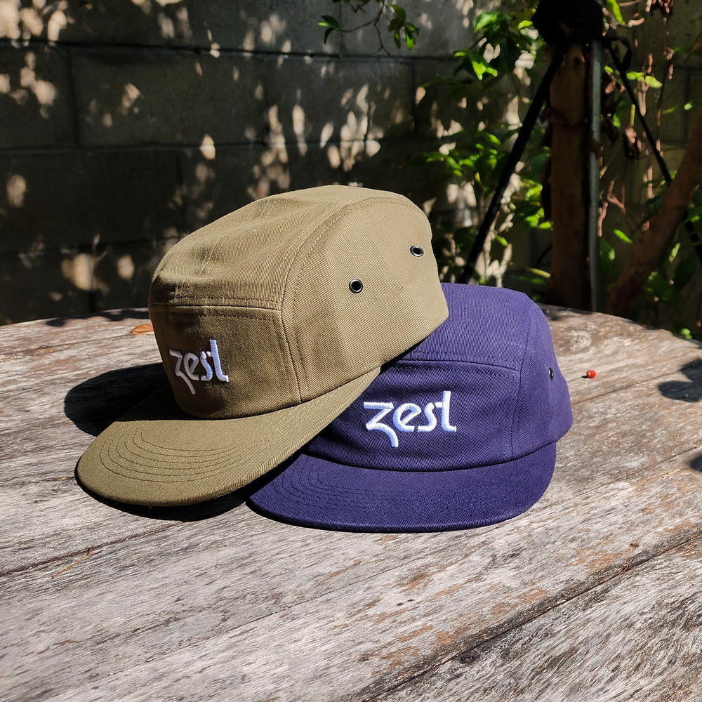 Olive and navy Zest five-panel caps stacked on wooden table outdoors in sunlight