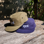 Olive and navy Zest five-panel caps stacked on wooden table outdoors in sunlight