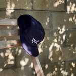 Navy Zest five-panel cap hanging on wooden post with sepia-toned brick wall background