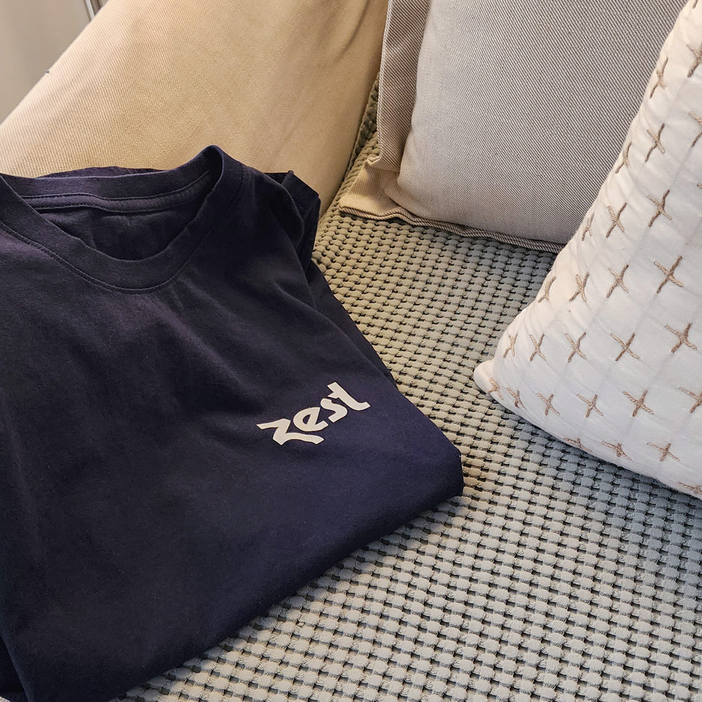 Folded navy Zest t-shirt on woven couch cushion with pillows