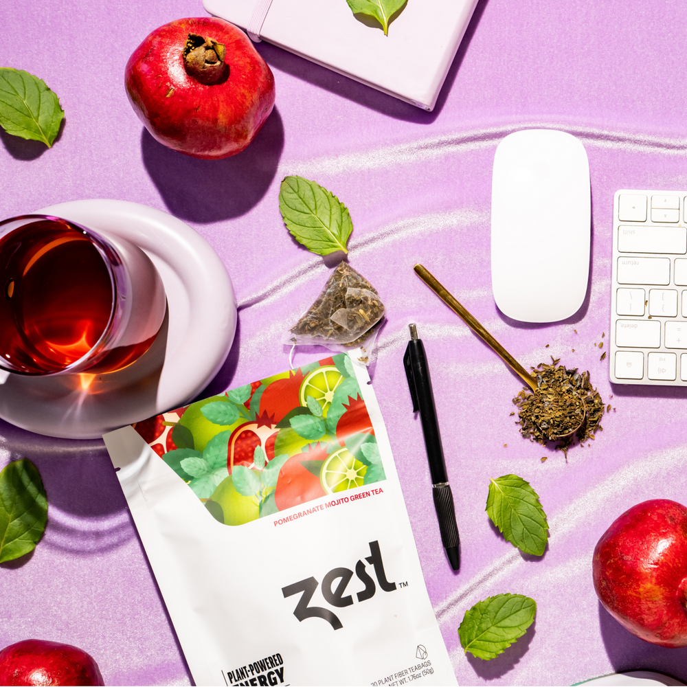 Zest Pomegranate Mojito pouch on purple desk with pomegranate, brewed tea, and keyboard